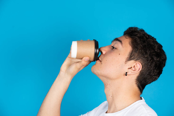 İspanyol genç adam mavi arka planda izole edilmiş tek kullanımlık ve geri dönüşümlü bardaktan kahve içiyor.. - Fotoğraf, Görsel