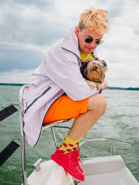 handsome blond young person wear raincoat sitting on a sailing yacht and hold his small dog yorkshire terrier during vacations - Фото, зображення