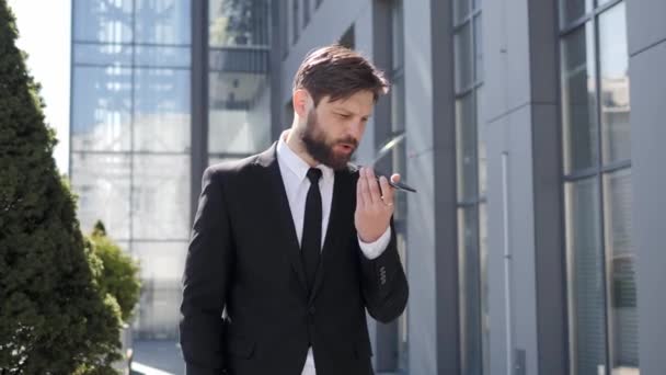 Stylish bearded man making voice message or using virtual assistant app on mobile phone. Confident businessman recording voice message on speakerphone while standing near modern office building. - Video