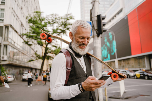 Grijze volwassen man met behulp van mobiele telefoon tijdens het staan met skateboard in de stad straat - Foto, afbeelding