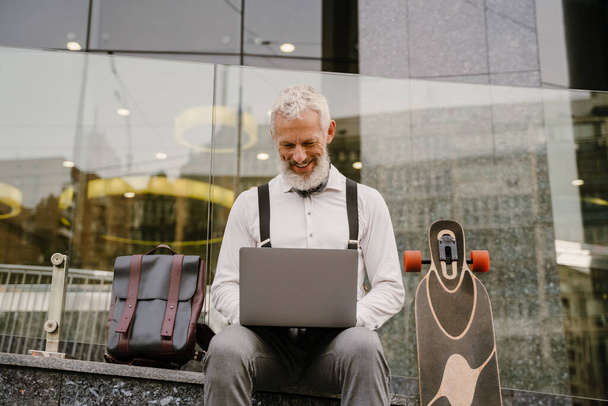 Grey mature man smiling while working with laptop while sitting at city street - 写真・画像