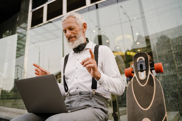 Grey mature man gesturing while working with laptop while sitting at city street - Foto, Bild