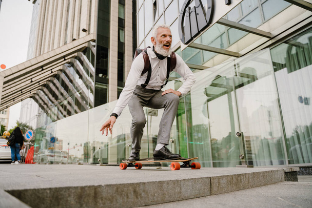 Grey mature man with beard smiling while skateboarding at city street - Photo, image