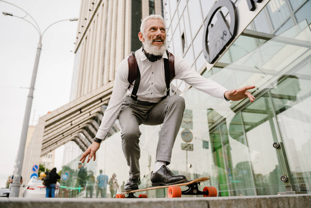 Grey mature man with beard smiling while skateboarding at city street - Фото, изображение