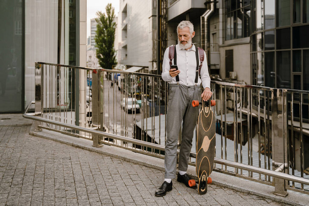 Grey mature man using cellphone while standing with skateboard at city street - Photo, Image