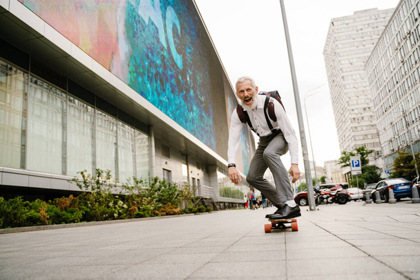 Grey mature man with beard smiling while skateboarding at city street - Foto, immagini
