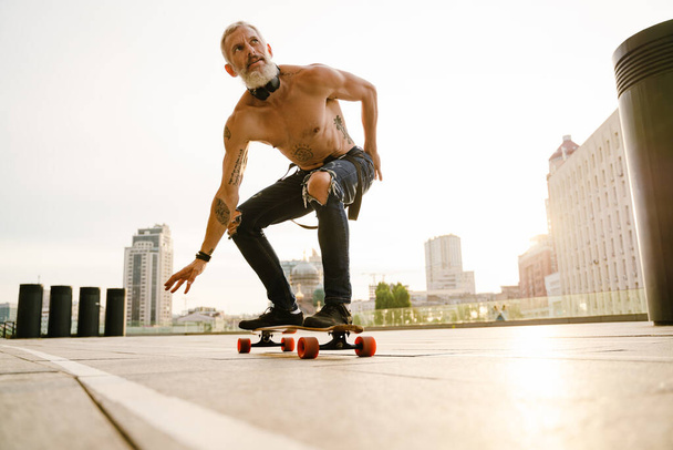 Shirtless mature man with beard skateboarding on parking outdoors - Photo, image