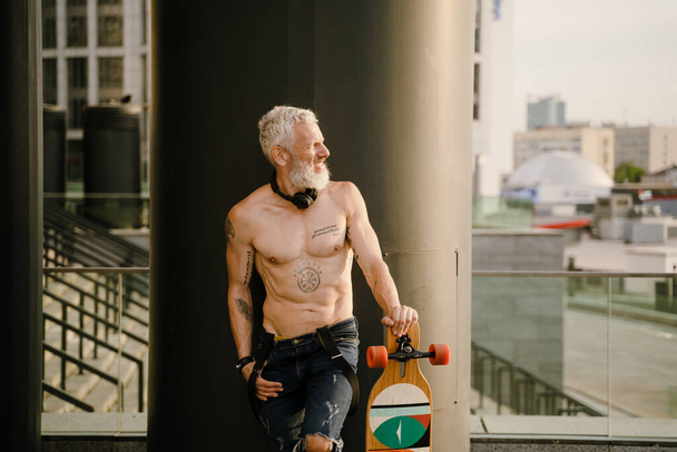 Shirtless mature man with tattoo standing with skateboard outdoors - Foto, afbeelding