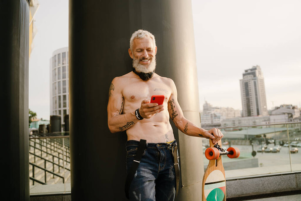 Shirtless mature man using cellphone while standing with skateboard at city street - Valokuva, kuva