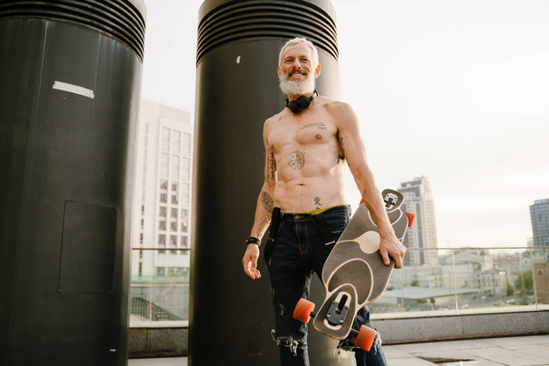 Взрослый мужчина без рубашки улыбается, стоя со скейтбордом на открытом воздухе - Фото, изображение
