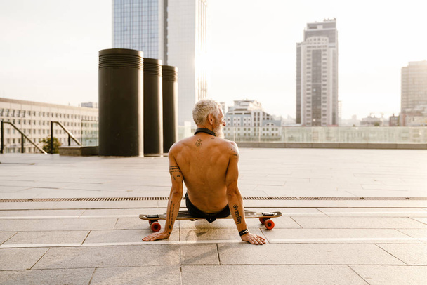 Shirtless mature man with tattoo sitting on skateboard outdoors - Foto, afbeelding