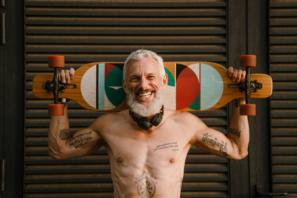 Shirtless mature man smiling while standing with skateboard outdoors - Foto, afbeelding