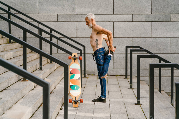 Shirtless mature man doing warmup while standing with skateboard on stairs outdoors - Fotó, kép