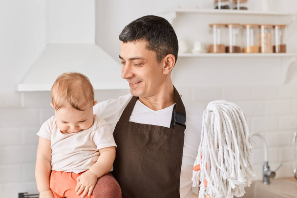 Portrait of Caucasian young adult father and infant baby daughter with floor mops in kitchen, man wearing brown apron, looking at child with happy facial expression. - Foto, Imagen