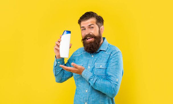 smiling brutal bearded man presenting hair conditioner bottle, presenting toiletries. - Fotografie, Obrázek