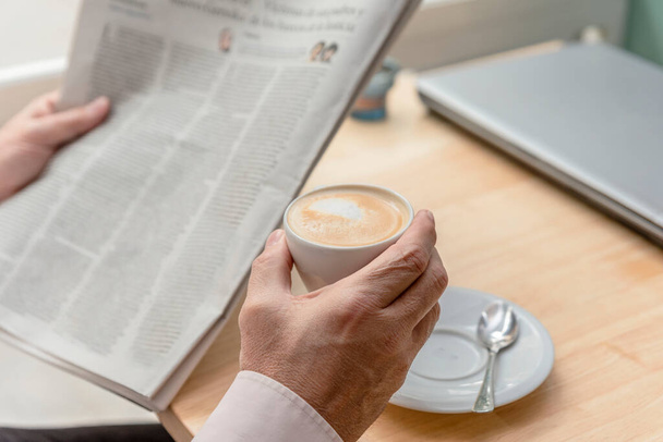 close-up of the hands of an unrecognizable older man holding a newspaper in one hand and a cup of coffee in the other, a closed laptop on the table, concept of leisure - 写真・画像