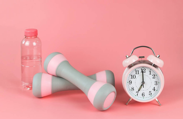 Front view of pink vintage alarm clock,  gray pink dumbbells and  bottle of water on pink background with copy space. Self development and active lifestyle. - Fotografie, Obrázek