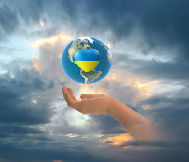 earth globe with Ukraine flag color blue and yellow in heart symbol in hands on front blue starry sky nebula hold world peace concept nature background - Photo, Image