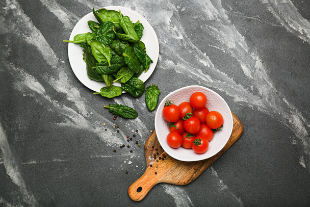 spinach leaves in a plate with tomatoes in a bowl on a wooden cutting board with salt and pepper. Black background. top view - Foto, Imagem