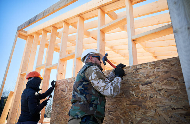 Carpenter hammering nail into OSB panel while building wooden frame house. - Fotoğraf, Görsel