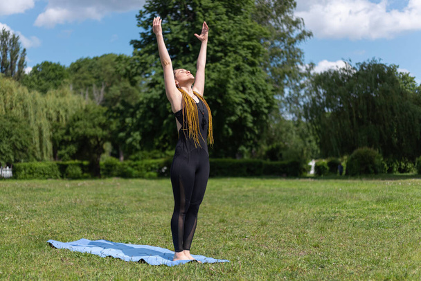 Woman yogi with dreadlocks stretching doing practicing Urdhva Hastasana upward hand pose on yoga mat in city park. Relaxing in the morning. Fitness instructor coach showing exercises  - Foto, imagen