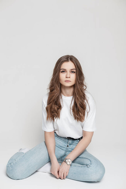 young pretty girl with clean skin and a hairstyle in a white T-shirt with fashionable blue jeans sits on a white background in the studio - Zdjęcie, obraz