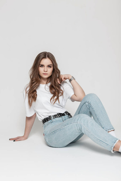 Glamorous beautiful serious girl model in casual clothes with a white t-shirt and blue high waist  jeans sits in the studio - Valokuva, kuva