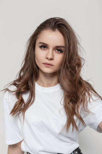 Pretty serious woman with cute face and hairstyle in white t-shirt look at the camera in studio. Fashion girl - 写真・画像