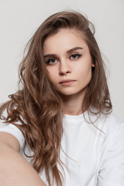 Κομψό νεαρό όμορφο κορίτσι με σγουρά μαλλιά και λευκό T-shirt σε λευκό φόντο στο στούντιο - Φωτογραφία, εικόνα