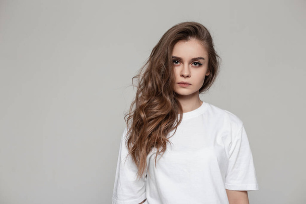 Female portrait of a beautiful girl with a hairstyle in a white T-shirt in the studio looks at the camera. Serious woman on gray background - Photo, image