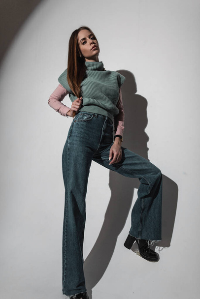 Сексуальна гламурна стильна дівчина в модному вінтажному одязі з в'язаним зеленим светром і рожевою сорочкою з модними джинсами дзвоника в студії. Красива жінка в ретро-стилі. Широкі джинси для ніг
 - Фото, зображення