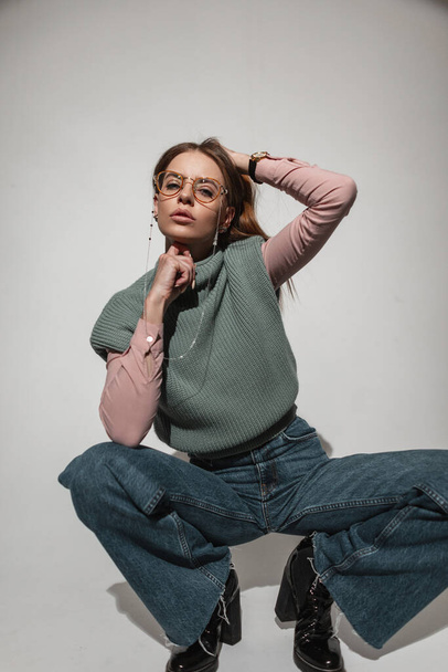 Stylish beautiful young girl model in fashionable vintage clothes with jeans, sweater and shirt with glasses sitting and posing in the studio with hard light - Foto, Imagen