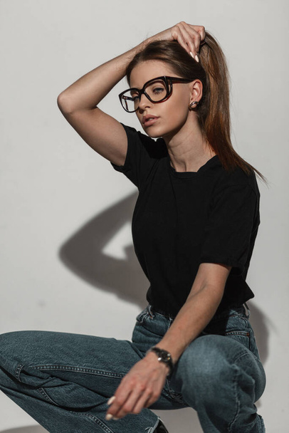 Pretty fashion woman with black glasses in stylish black t-shirt mock-up and jeans sits in studio. Cool girl poses on a white background - Foto, afbeelding