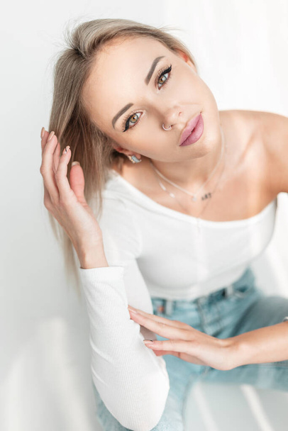 Beautiful young stylish blonde woman with green eyes in a white T-shirt and blue jeans sits in a studio with sunlight - Photo, Image