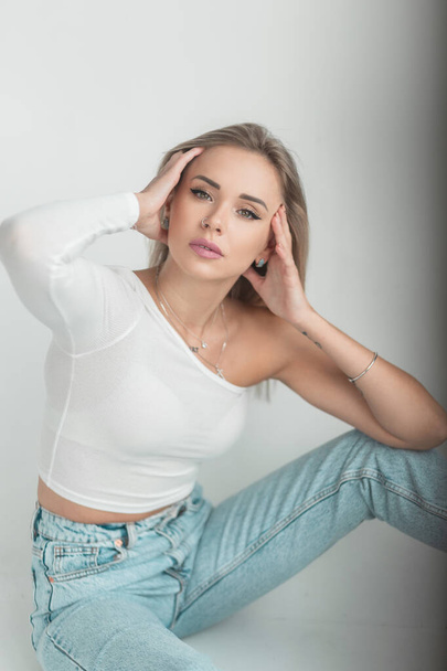 Cute stylish woman blonde model in white t-shirt and vintage blue jeans sits on white floor in studio - Fotó, kép