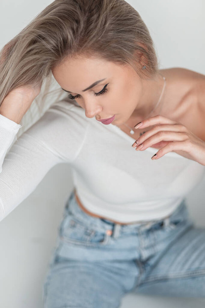 Beautifully young blonde woman model with pretty female face in white t-shirt and blue vintage jeans sits on a white background - Fotoğraf, Görsel