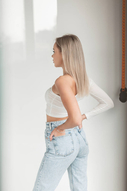 Stylish beautiful young blonde woman in fashionable clothes with white t-shirt and vintage blue jeans in the studio with highlights effect - Fotoğraf, Görsel