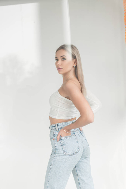 Beautiful blonde girl in a fashionable white T-shirt and blue vintage high waist jeans in the studio on a white background with glare effects - 写真・画像