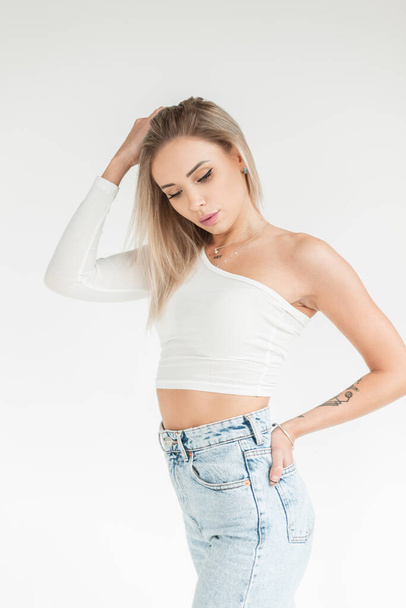 Stylish beautiful young blonde woman with a cute face in a fashionable white t-shirt with blue high waist jeans on a white background - Fotó, kép