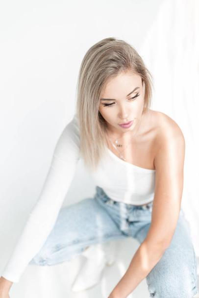 Delicate beautiful young blonde woman with a white t-shirt and vintage blue jeans sits on a white background in sunlight - Photo, Image