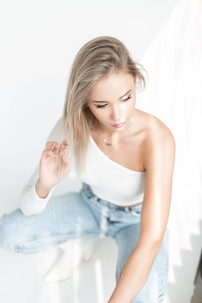 Красива молода модель блондинки в моді біла футболка і блакитні джинси сидить на білому тлі на сонячному світлі
 - Фото, зображення