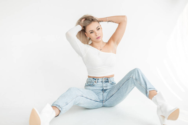 Fashionable beautiful young blonde woman in a stylish white tank top with vintage blue jeans and shoes sits on a white background in a studio with sunlight - Fotografie, Obrázek