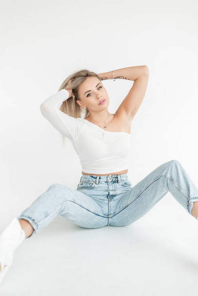 jong mooi blond meisje in een modieus wit T-shirt met blauwe vintage jeans zit op een witte achtergrond in de studio - Foto, afbeelding
