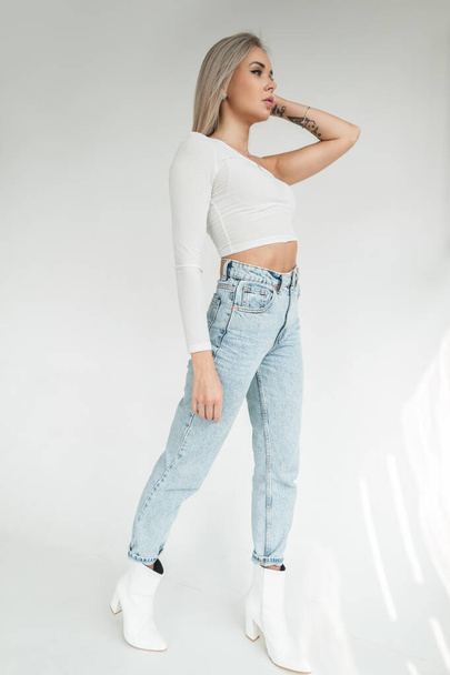 Stylish beautiful gentle blonde girl in a fashionable white T-shirt and blue vintage high waist jeans with white shoes on a white background in the studio - 写真・画像