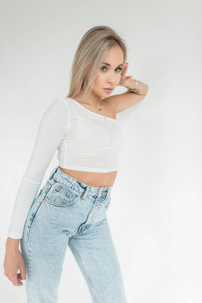 Menina loira muito jovem em uma camiseta branca e jeans de cintura alta azul em um fundo branco no estúdio olha para a câmera - Foto, Imagem