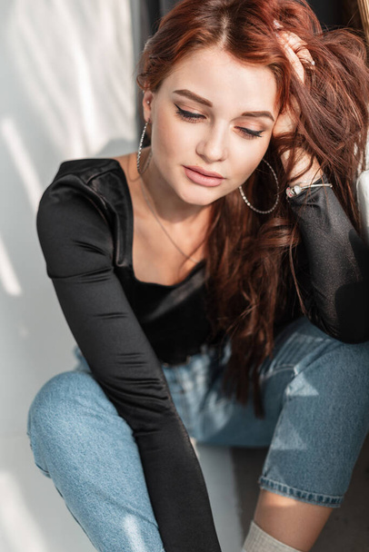 Female portrait of a beautiful young stylish woman with a cute face in a black t-shirt with blue jeans sits on the floor in the rays of the sun - Foto, Bild