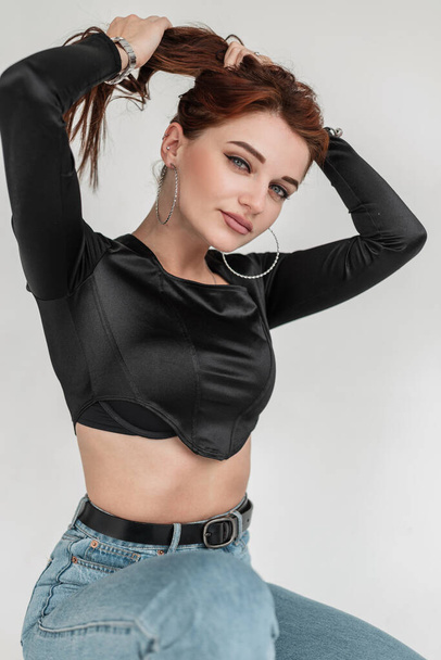 Stylish glamorous young woman in trendy black clothes with blue jeans in studio - Фото, зображення