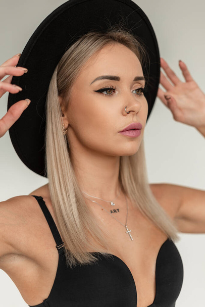 Sexy portrait of beautiful young blonde woman with stylish hat and fashionable black bra poses in studio - Foto, afbeelding