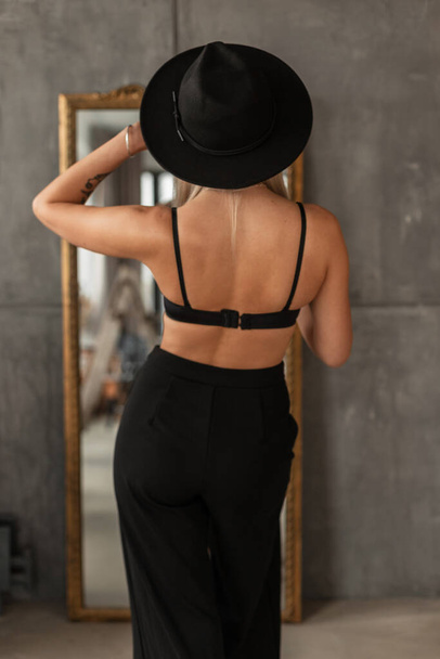 Beautiful elegant woman in a fashionable black hat with stylish pants and a black wall lamp stands near the mirror with her back to the camera - Valokuva, kuva