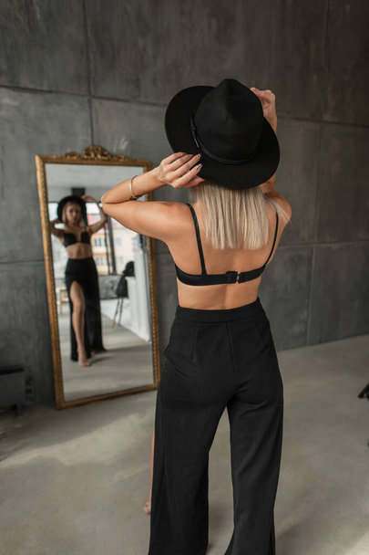 Stylish beautiful young blonde woman with a vintage hat in fashionable clothes with a bra and trousers stands near the mirror in the room - Foto, Bild